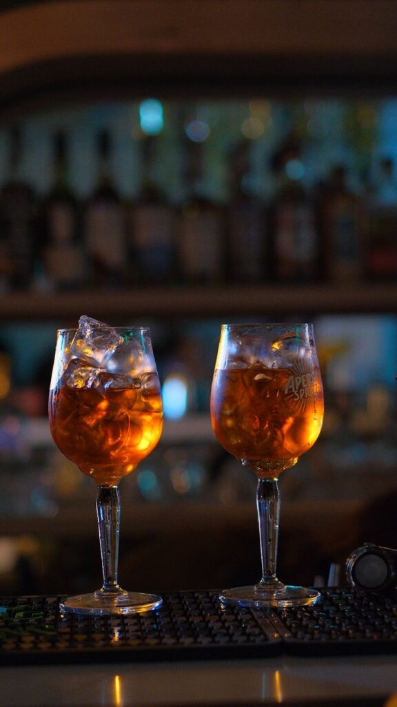 Two Aperol Spritz cocktails with ice cubes on a dimly lit bar counter.